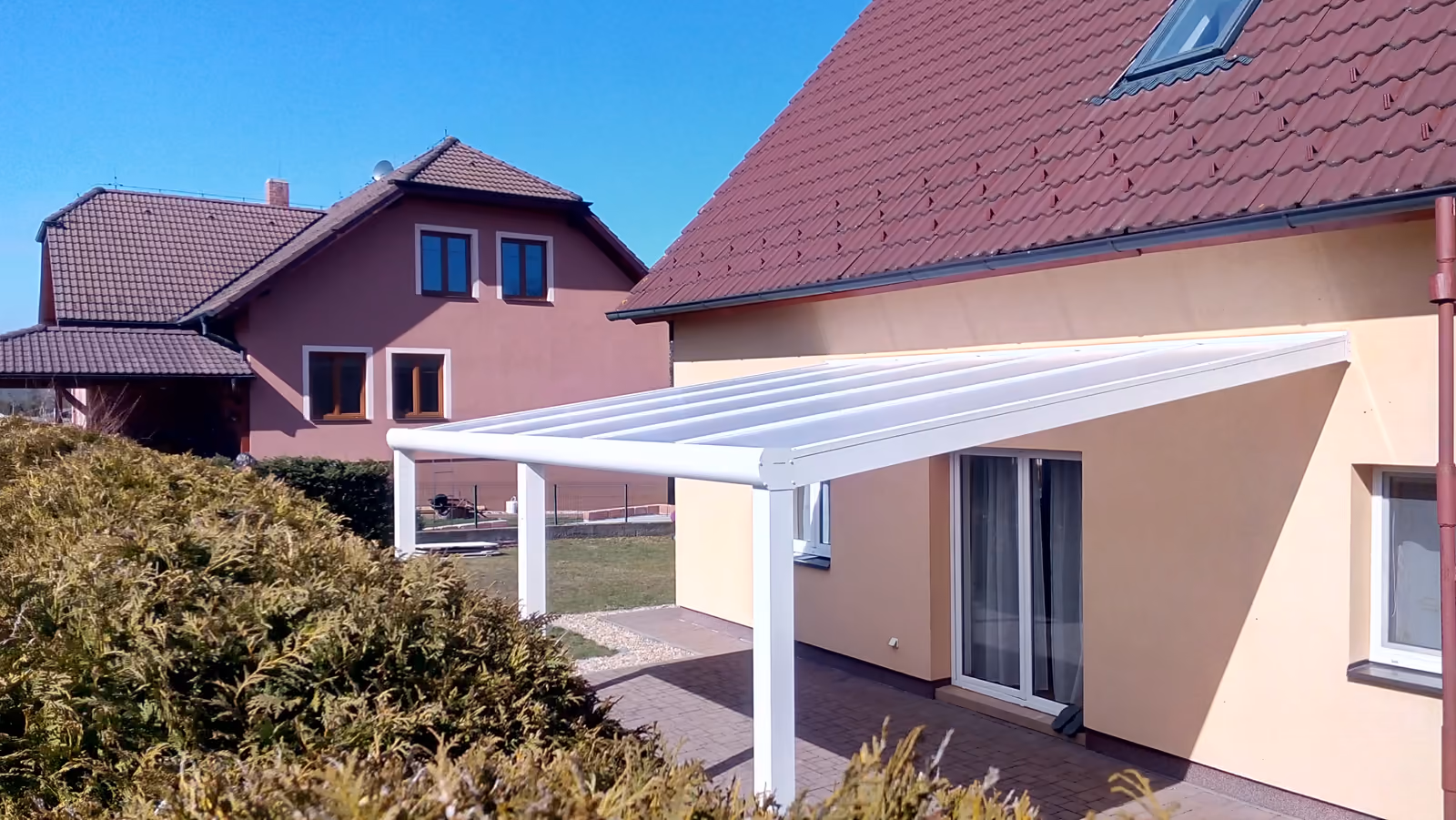 bílá hliníková pergola nad dlážděnou terasou u béžového domu