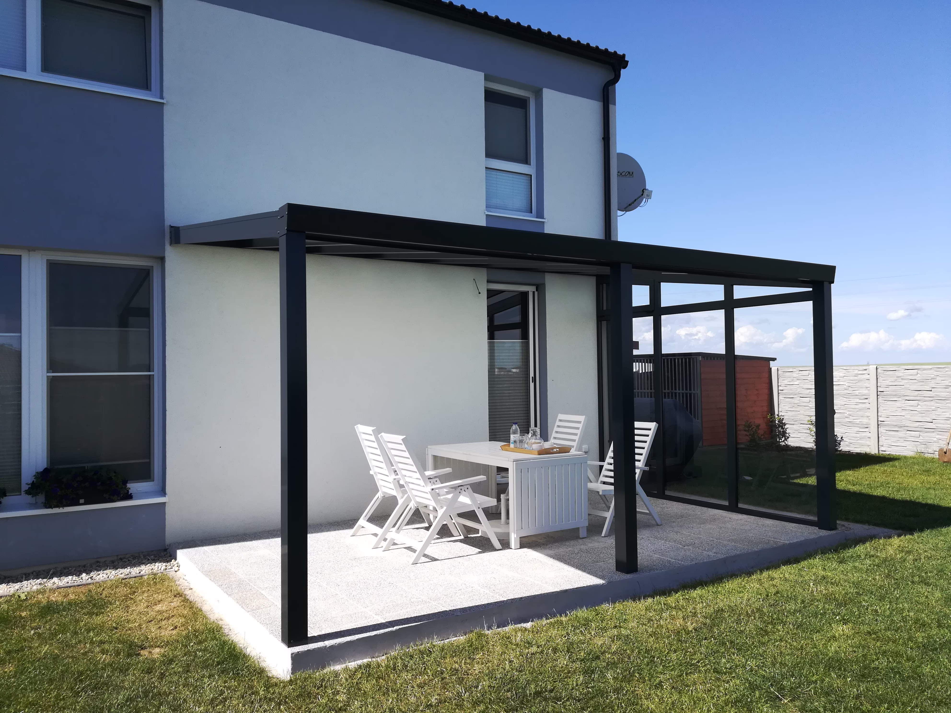 antracitová pergola se skleněnou stěnou nad terasou u bílého domu
