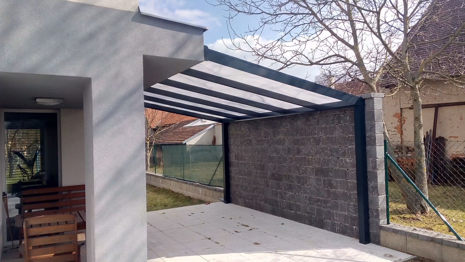 antracitová pergola nad šedou dlažbou vedle kamenné stěny